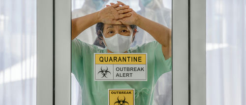A healthcare worker in a green uniform and mask, looking distressed behind a glass door marked with quarantine signs