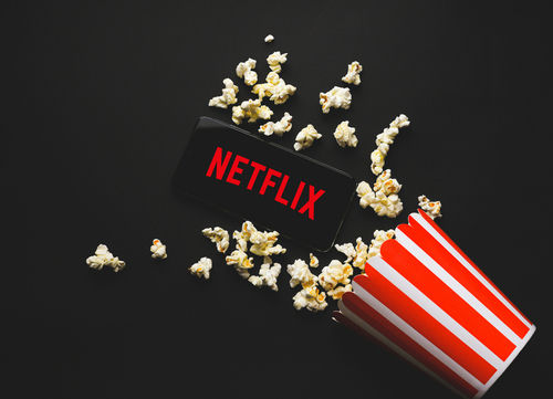 shutterstock_1828714433.jpg Spilled popcorn, phone displaying Netflix logo, striped popcorn container.