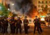 Police officers in riot gear facing a chaotic protest scene with smoke and fire