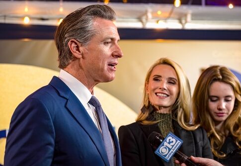 A man in a suit speaking at an event with two women nearby, one smiling and the other holding a microphone
